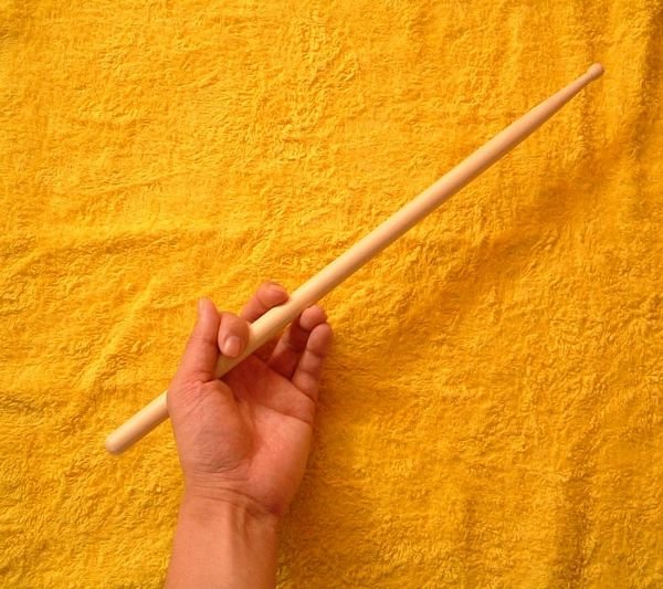 A hand holding a drumstick. The thumb and index finger pinch the stick, the other three fingers curl under it, and a short length of the butt shows behind the hand.